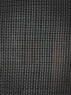 A tightly woven metal grid pattern with small rectangular openings. The background is dark, slightly visible through the mesh.