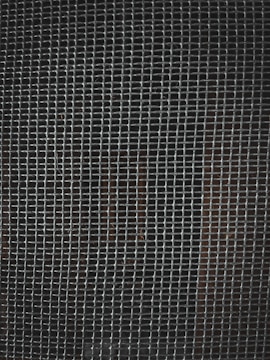 A tightly woven metal grid pattern with small rectangular openings. The background is dark, slightly visible through the mesh.