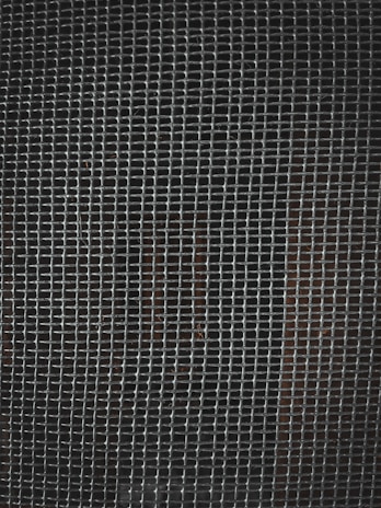 A tightly woven metal grid pattern with small rectangular openings. The background is dark, slightly visible through the mesh.