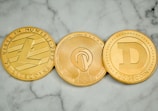 Three round, metallic coins with distinct engravings and symbols are placed side by side on a marble surface. The left coin has an 'L' logo and the word 'LITECOIN' inscribed around its edge. The middle coin features a more intricate design with circuit-like patterns. The right coin displays a large letter 'D' and the word 'DOGECOIN' around its perimeter.