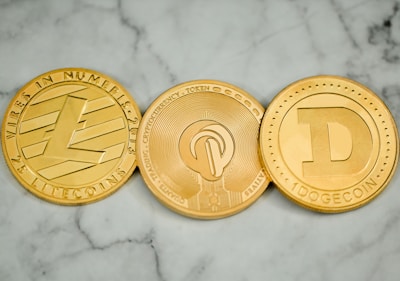 Three round, metallic coins with distinct engravings and symbols are placed side by side on a marble surface. The left coin has an 'L' logo and the word 'LITECOIN' inscribed around its edge. The middle coin features a more intricate design with circuit-like patterns. The right coin displays a large letter 'D' and the word 'DOGECOIN' around its perimeter.