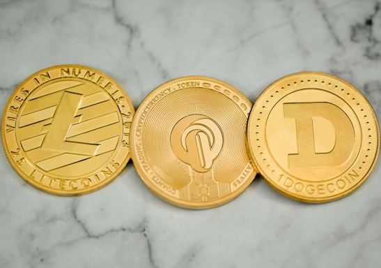 Three round, metallic coins with distinct engravings and symbols are placed side by side on a marble surface. The left coin has an 'L' logo and the word 'LITECOIN' inscribed around its edge. The middle coin features a more intricate design with circuit-like patterns. The right coin displays a large letter 'D' and the word 'DOGECOIN' around its perimeter.