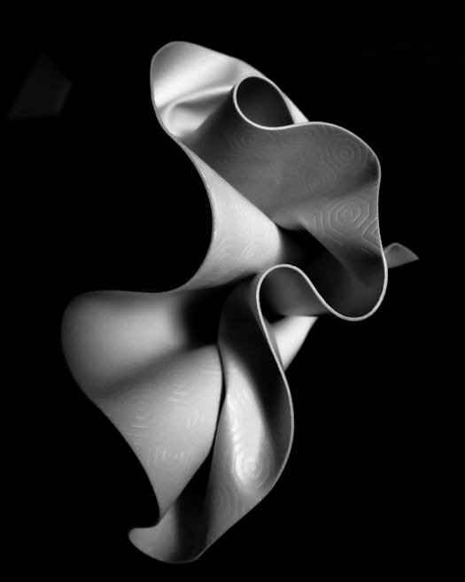 A flowing, abstract sculpture with smooth, curved surfaces that resemble intricate waves or folds. The surface displays subtle geometric patterns, adding texture to the form, and it is set against a deep black background, enhancing the contrast.