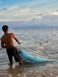 A person standing in shallow water, pulling a large fishing net filled with shells or small floats. The ocean extends into the horizon under a partly cloudy sky with hues of blue and a hint of yellow from the setting or rising sun.