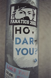 A weathered sticker on a metal pole displays a stylized face with the word 'FANATICS 2011.' Below, partially torn text reads 'HO DAR YOU?' in large lettering. Additional text and graphics are underneath, partly obscured.
