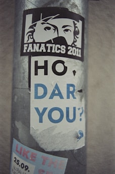 A weathered sticker on a metal pole displays a stylized face with the word 'FANATICS 2011.' Below, partially torn text reads 'HO DAR YOU?' in large lettering. Additional text and graphics are underneath, partly obscured.