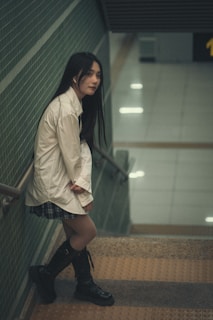 A woman with long dark hair is leaning against the railing on a staircase. She is wearing a white shirt, a plaid skirt, and knee-high black boots. The setting appears to be an indoor location with green-tiled walls and yellow tactile paving on the steps.