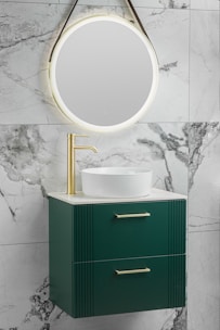 A modern bathroom vanity setup featuring a round, illuminated mirror mounted on a marble wall. Below the mirror is a sleek green cabinet with two drawers, each fitted with gold handles. A white vessel sink sits on top of the cabinet, paired with a gold faucet.