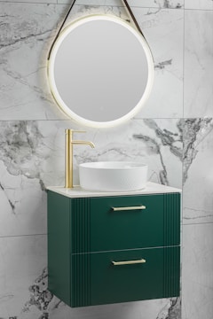 A modern bathroom vanity setup featuring a round, illuminated mirror mounted on a marble wall. Below the mirror is a sleek green cabinet with two drawers, each fitted with gold handles. A white vessel sink sits on top of the cabinet, paired with a gold faucet.