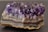 A large, rough-textured amethyst geode with vivid purple crystals protruding from a layered stone base. The crystals are sharp and translucent, catching the light to reveal various shades of violet and lavender, while the base exhibits earthy tones of brown, grey, and white.