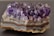 A large, rough-textured amethyst geode with vivid purple crystals protruding from a layered stone base. The crystals are sharp and translucent, catching the light to reveal various shades of violet and lavender, while the base exhibits earthy tones of brown, grey, and white.