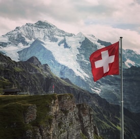 A scenic landscape featuring a majestic mountain range with snow-capped peaks. A Swiss flag waves prominently in the foreground, attached to a pole on a rocky cliff covered with green vegetation. The sky is overcast, adding to the dramatic effect of the scene.