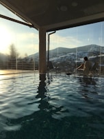 An indoor pool reflecting sunlight, with large glass windows framing a view of snowy mountains. A person sits on a lounge chair near the window, enjoying the serene ambiance.