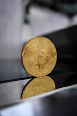 A gold Bitcoin coin is standing upright on a reflective surface, with a blurred background. The bottom of the coin is visible along with its reflection, highlighting the intricate details and the Bitcoin symbol on its face.