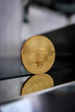 A gold Bitcoin coin is standing upright on a reflective surface, with a blurred background. The bottom of the coin is visible along with its reflection, highlighting the intricate details and the Bitcoin symbol on its face.