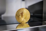 A gold-colored Bitcoin coin is placed upright on a reflective surface, showcasing its digital design and markings. The background is blurred, which emphasizes the coin's intricate details and symbolizes cryptocurrency.
