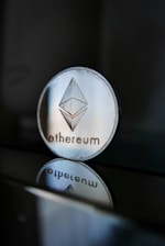 A close-up of an Ethereum coin with its logo clearly visible, placed on a glossy surface that reflects the coin's image beneath it. The background is dark, creating a stark contrast with the shiny metallic appearance of the coin.