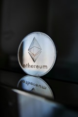 A close-up of an Ethereum coin with its logo clearly visible, placed on a glossy surface that reflects the coin's image beneath it. The background is dark, creating a stark contrast with the shiny metallic appearance of the coin.