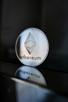 A close-up of an Ethereum coin with its logo clearly visible, placed on a glossy surface that reflects the coin's image beneath it. The background is dark, creating a stark contrast with the shiny metallic appearance of the coin.