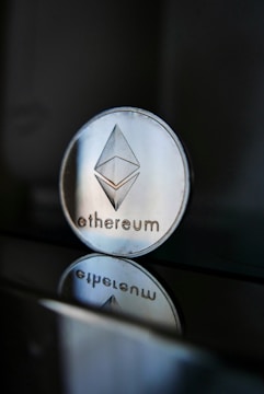 A close-up of an Ethereum coin with its logo clearly visible, placed on a glossy surface that reflects the coin's image beneath it. The background is dark, creating a stark contrast with the shiny metallic appearance of the coin.