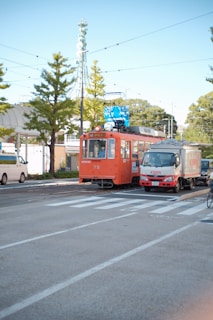 A vibrant orange tram is positioned on a city street next to a white and red delivery truck displaying the brand 'KIRIN'. Overhead wires and traffic lights are visible, with green trees lining the street. Other vehicles, including a car and a cyclist, are present near the pedestrian crossing.