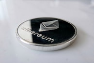 A close-up view of a metallic coin featuring the Ethereum logo and the word 'ethereum' inscribed on it. The coin has a silver rim and dark center, lying on a smooth white surface.