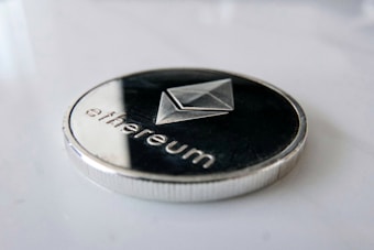 A close-up view of a metallic coin featuring the Ethereum logo and the word 'ethereum' inscribed on it. The coin has a silver rim and dark center, lying on a smooth white surface.