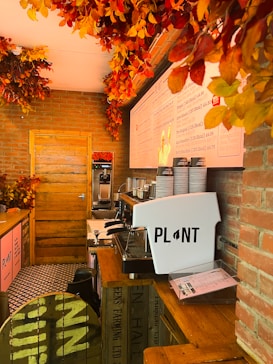 Interior of a cozy café with brick walls, decorated with autumn leaves on the ceiling. A wooden counter features a large coffee machine labeled 'PLANT' and stacks of paper cups. Menus and decorations hang on the walls, contributing to a warm, welcoming atmosphere.