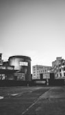 An urban environment featuring industrial buildings with a prominent cylindrical structure in the foreground displaying text related to an exhibition. Surrounding structures have an unfinished or minimalistic appearance, and the area appears vacant.
