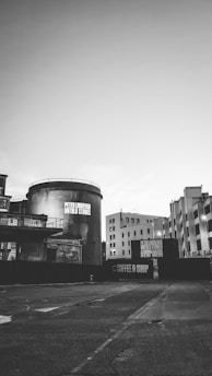 An urban environment featuring industrial buildings with a prominent cylindrical structure in the foreground displaying text related to an exhibition. Surrounding structures have an unfinished or minimalistic appearance, and the area appears vacant.