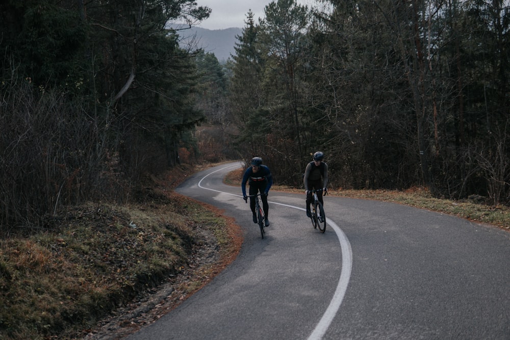 two bicyclists riding down a winding road in the woods