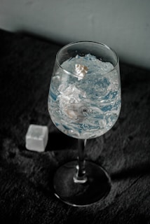 A wine glass filled with clear ice cubes stands on a dark textured surface. The light reflects off the ice, creating a sparkling effect. In the background, a single ice cube lies on the surface near the glass.