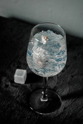 A wine glass filled with clear ice cubes stands on a dark textured surface. The light reflects off the ice, creating a sparkling effect. In the background, a single ice cube lies on the surface near the glass.