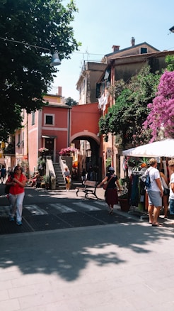A charming street scene in a European village, featuring a mix of rustic buildings with vibrant colors. Bougainvillea flowers cascade over the walls, and a few people are walking along the stone pavement, exploring small shops and enjoying the leisurely atmosphere. An arched doorway leads to a staircase, enhancing the picturesque setting.