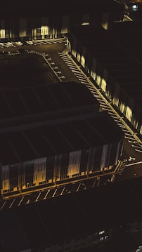 An aerial view of a dimly lit parking area, adjacent to large industrial or commercial buildings. The structures appear to have a modern architectural design, with angular lines and illuminated facades. Streetlights cast a warm glow on the parking spaces and driveways, creating a contrast between the dark shadows and the light.