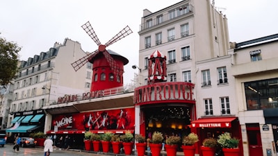 A lively street scene featuring the iconic red windmill and entrance of a famous cabaret venue. The building is adorned with large red signs and decorative elements, surrounded by potted plants in bright red containers. The venue is set against a backdrop of typical Parisian architecture, with multi-story buildings and shops lining the street. A few pedestrians and vehicles can be seen on the road.