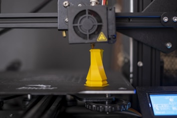 A 3D printer is in the process of creating a yellow chess piece, possibly a rook. The printer consists of a mechanical arm with a nozzle extruding yellow filament material onto the printing surface. The background shows the printer's structure, including its frame and supporting components.