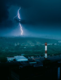 A powerful lightning bolt strikes down from a dark, stormy sky over an industrial area with a tall, striped smokestack in the foreground. The cityscape is partially visible under the storm's heavy clouds.