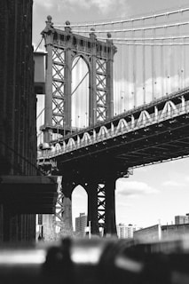 A black and white photograph of a large suspension bridge with intricate metalwork. The bridge towers over urban buildings and is adorned with cables stretching into the sky. The foreground has a slightly blurred silhouette of people, adding a sense of scale and life to the urban scene. The architecture is detailed and industrial, with the skyline visible in the background.