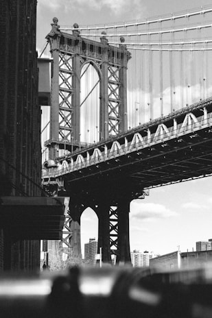 A black and white photograph of a large suspension bridge with intricate metalwork. The bridge towers over urban buildings and is adorned with cables stretching into the sky. The foreground has a slightly blurred silhouette of people, adding a sense of scale and life to the urban scene. The architecture is detailed and industrial, with the skyline visible in the background.