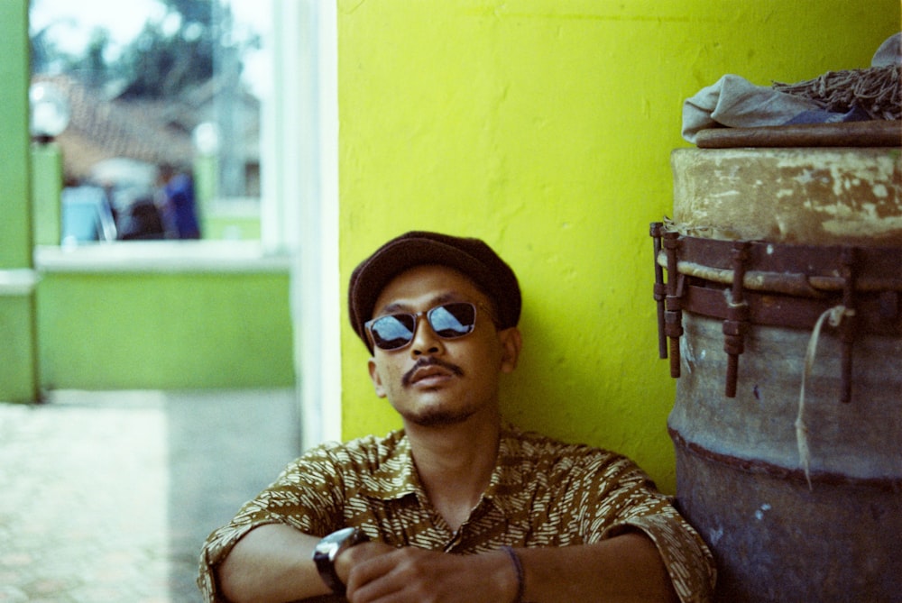 a man wearing sunglasses leaning against a yellow wall