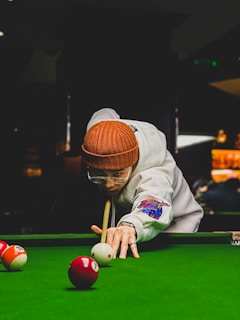 A person is concentrating on making a shot in a game of billiards. They are wearing a brown knitted hat, a white jacket with colorful patches, and glasses. The person is leaning over the pool table, focusing intently on a cue ball as they hold a pool cue in position. The table is covered with a vibrant green felt, and several billiard balls are scattered around.