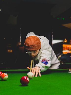 A person is concentrating on making a shot in a game of billiards. They are wearing a brown knitted hat, a white jacket with colorful patches, and glasses. The person is leaning over the pool table, focusing intently on a cue ball as they hold a pool cue in position. The table is covered with a vibrant green felt, and several billiard balls are scattered around.
