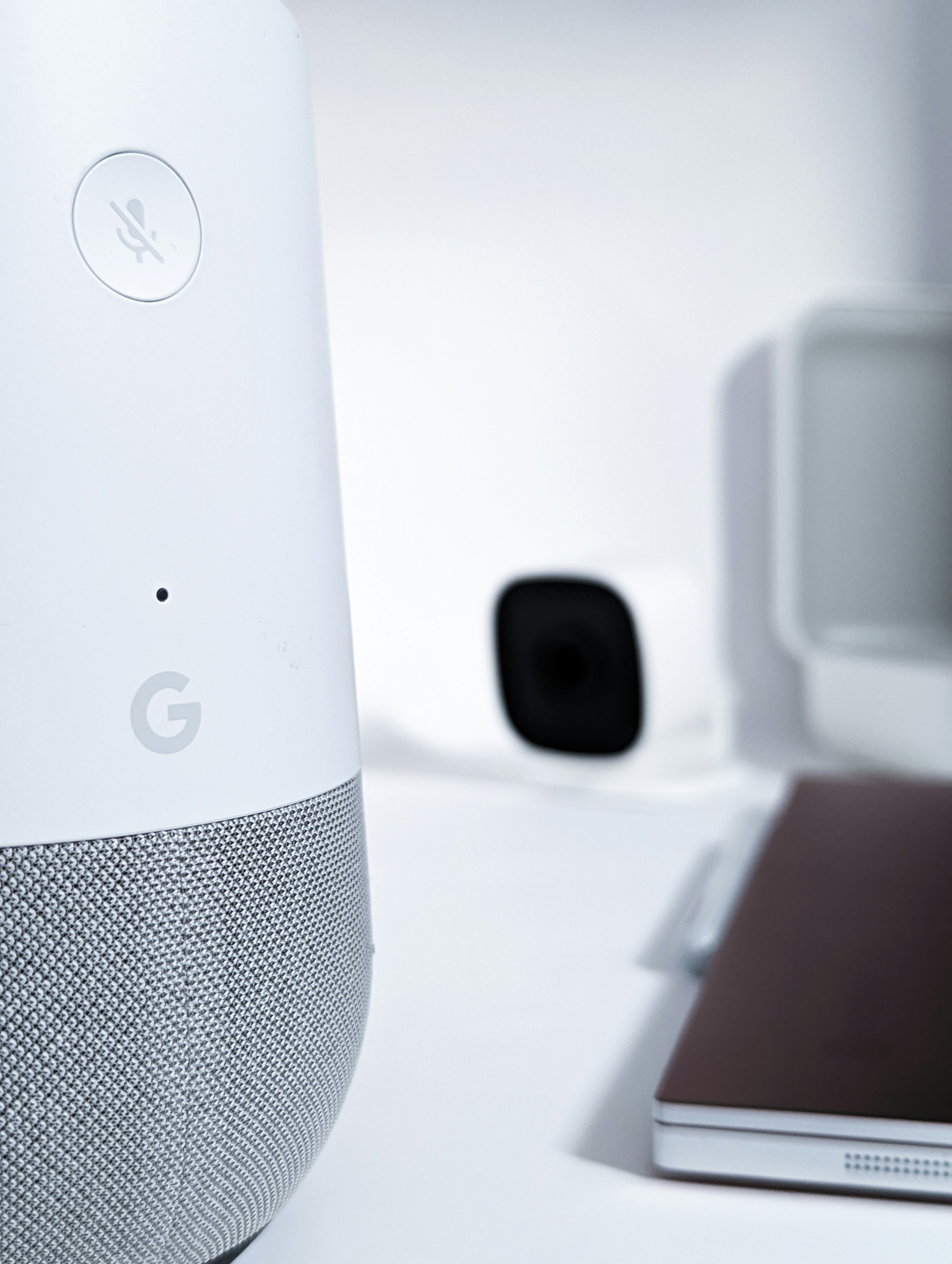 A close-up view of a smart speaker with a mute button, featuring a sleek design in white and gray. In the background, there is a blurry image of a camera and a laptop, creating a high-tech and minimalist workspace environment.