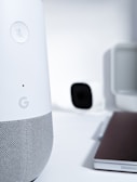 A close-up view of a smart speaker with a mute button, featuring a sleek design in white and gray. In the background, there is a blurry image of a camera and a laptop, creating a high-tech and minimalist workspace environment.