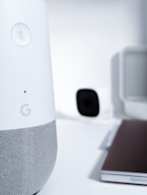A close-up view of a smart speaker with a mute button, featuring a sleek design in white and gray. In the background, there is a blurry image of a camera and a laptop, creating a high-tech and minimalist workspace environment.