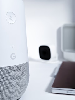 A close-up view of a smart speaker with a mute button, featuring a sleek design in white and gray. In the background, there is a blurry image of a camera and a laptop, creating a high-tech and minimalist workspace environment.