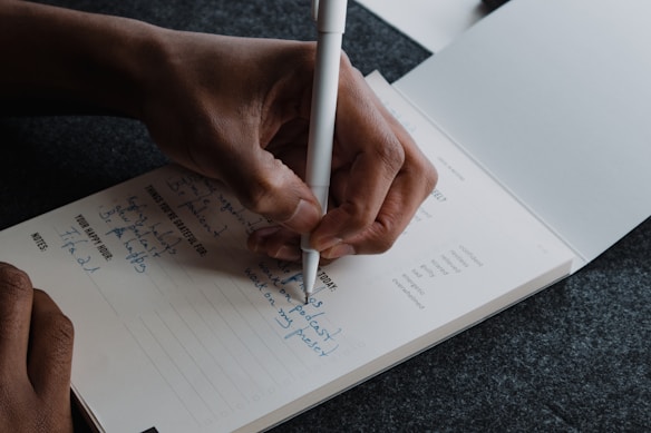 A person is writing in a notebook with a white pen. The notebook contains handwritten notes in blue ink, some of which list things to be grateful for, alongside a section titled 'Your Happy Hour'. The person's hand is positioned above the notebook, holding the pen.