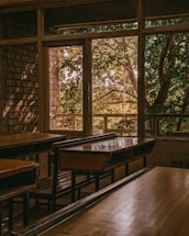 An empty classroom with wooden desks and benches is dimly lit by natural light coming through large windows. Outside, lush green trees are visible, creating a contrast with the warm tones inside.