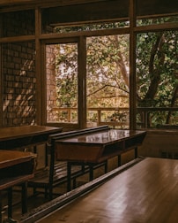 An empty classroom with wooden desks and benches is dimly lit by natural light coming through large windows. Outside, lush green trees are visible, creating a contrast with the warm tones inside.